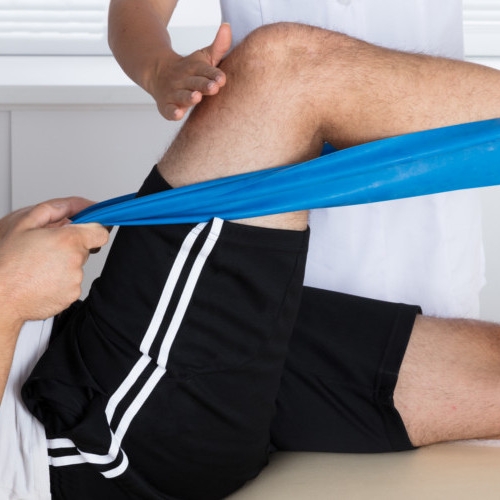 Male patient receiving physical therapy on his knee aided by a physical therapist and using resistance band.